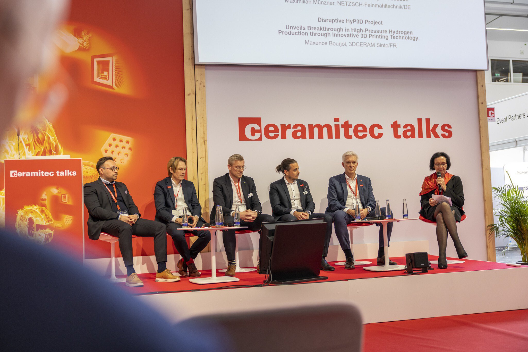 ceramitec Talks with six speakers on stage in front of a red background, a woman speaks into a microphone.