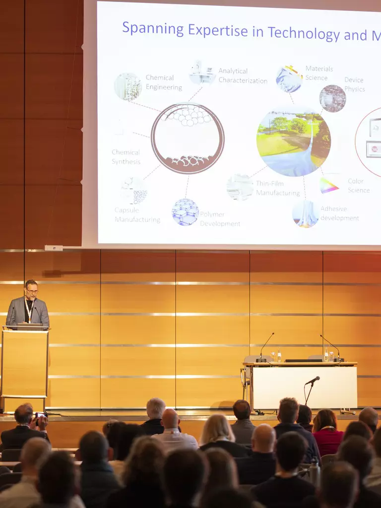 A speaker gives a presentation at the LOPEC conference in a large room and shows a slide with various diagrams to an audience.