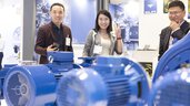 Three smiling people, in front of them large blue electric motors are displayed at an exhibition stand.