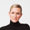 A woman with blonde hair, wearing a black turtleneck jumper, is sitting on a white chair and smiling at the camera.