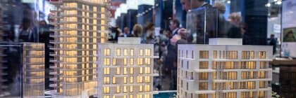 View of an architectural model in a glass display case with three illuminated high-rise buildings. Numerous visitors can be seen behind it.