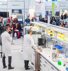 Cameramen film a woman in a lab coat at an exhibition stand. People follow the demonstration in the background.