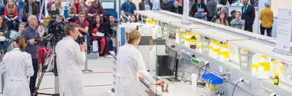 Kameraleute filmen an einem Messestand eine Frau in Laborkittel. Im Hintergrund folgen Personen der Vorführung.