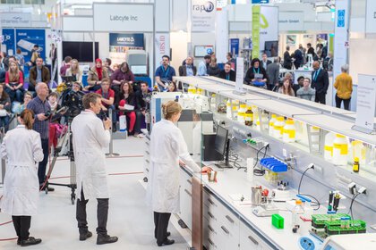Cameramen film a woman in a lab coat at an exhibition stand. People follow the demonstration in the background.