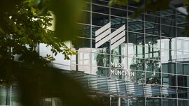 Gläserner Eingang der Messe München mit Logo, teilweise eingerahmt von grünen Blättern.