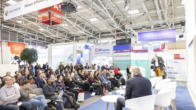 Power Efficiency Stage at electronica. One speaker is standing with a microphone, the other is sitting at a table on the stage. In front of them are full rows of spectators.
