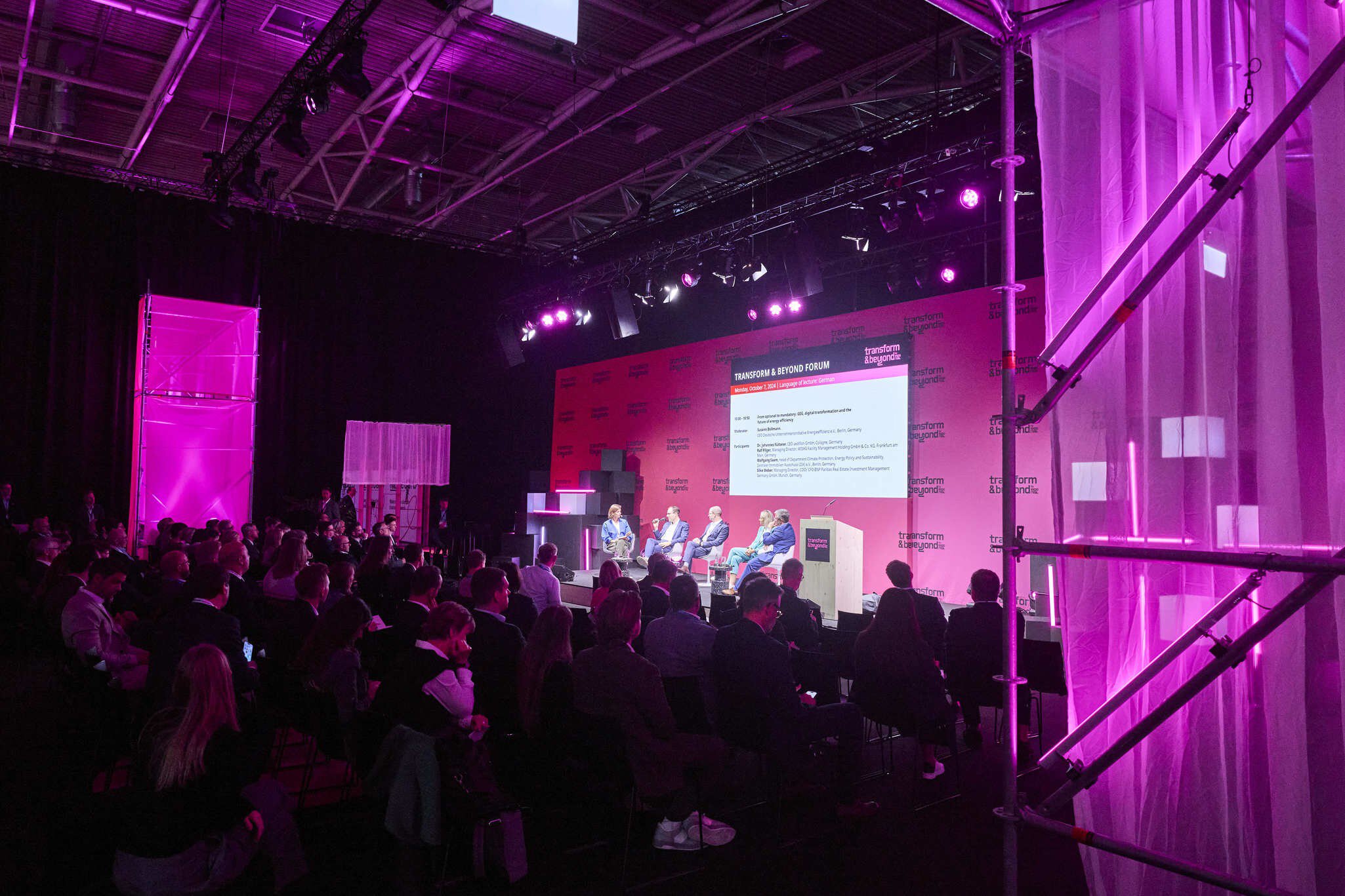 The audience is watching a panel discussion between 5 people on the stage of the transform & beyond forum. The room in front of the stage is dimly lit in violet.