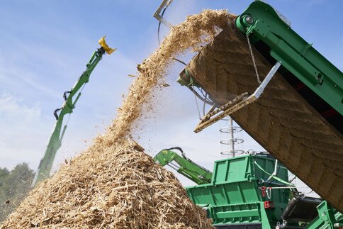 In the open-air exhibition area, a conveyor belt boom of a machine transports wood chips onto a large pile.