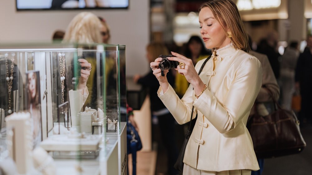 A woman with long, reddish hair is wearing a beige designer suit and is taking pictures of several pieces of jewelry in a display case with a camera.