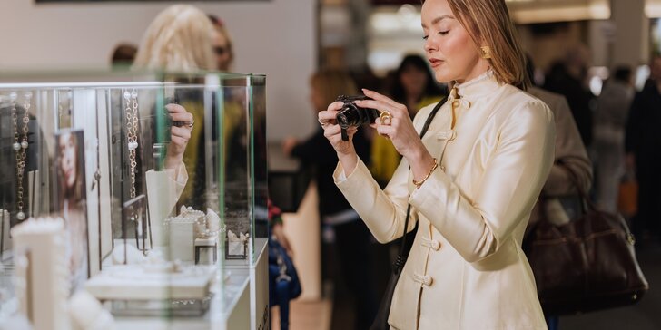 A woman with long, reddish hair is wearing a beige designer suit and is taking pictures of several pieces of jewelry in a display case with a camera.