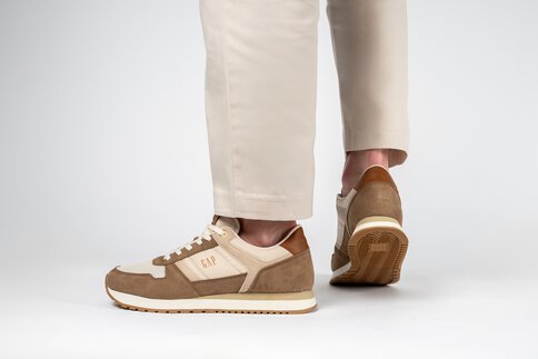 Ein Paar Beine in weißer Hose stecken in braun-beigen Halbschuhen der Marke „GAP“.