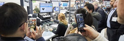 Eine Frau sitzt an einem Stand auf der productronica, während hinter ihr Zuschauer die Demonstration ansehen und filmen.