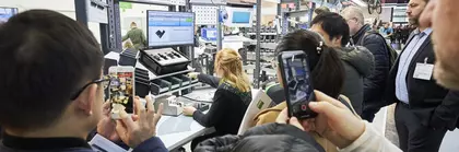 A woman sits at a stand at productronica while spectators watch and film the demonstration behind her.