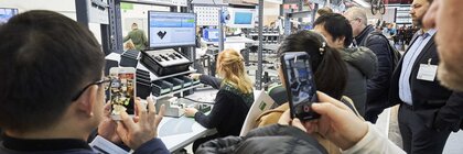 A woman sits at an ergonomically optimized workstation while viewers watch and film her work behind her.