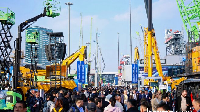 Many people walk through an outdoor exhibition area with construction machines, cranes, and information signs.