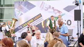 Menschen sitzen mit Getränken an Tischen im Freien und unterhalten sich. Im Hintergrund spielt eine Blaskapelle auf einer Bühne.