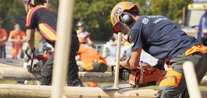 Zwei Männer in Sicherheitsausrüstung fräsen mit Kettensägen auf der INTERFORST im Freien lange Holzstücke ab, die in liegende Baumstämme gesteckt wurden.