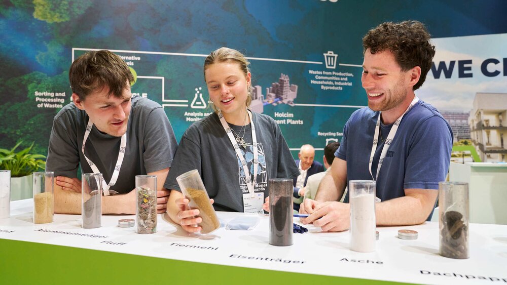 Three young trade fair visitors examining various raw material samples, such as ash and iron carriers, presented in transparent tubes on a counter.