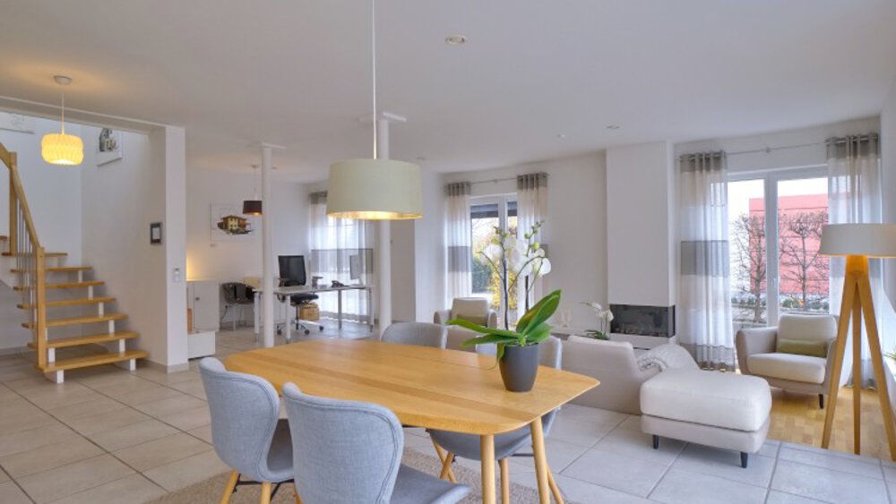 Moderner offener Wohnraum in neutralen Tönen, mit Esstisch, Stühlen, großen Fenstern und Treppe.