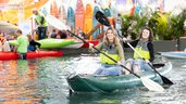 Two women sit in an inflatable kayak on a body of water inside the exhibition hall and paddle side by side. In the background, more boats, paddlers, and exhibitor stands can be seen.