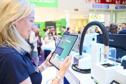 A woman stands at a trade fair booth holding a tablet. In the background, modern laboratory equipment and several visitors exploring the exhibition can be seen.