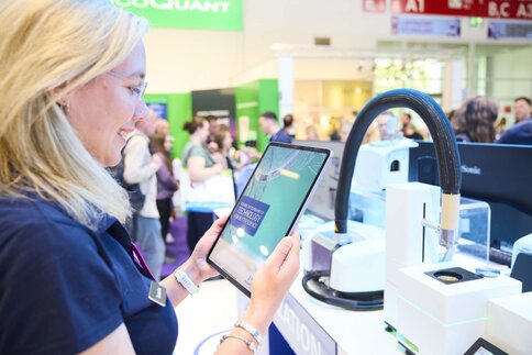 Eine Frau steht an einem Messestand und hält ein Tablet in den Händen. Im Hintergrund befinden sich moderne Laborgeräte und mehrere Messebesucher