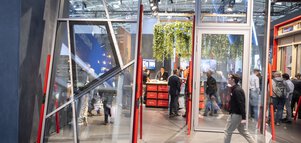 Metallrahmen mit Glasfenstern und Türen in gewöhnlichen und auch unregelmäßgen Formen stehen ausgestellt an einem Messestand. Dazwischen zahlreiche Besucher.