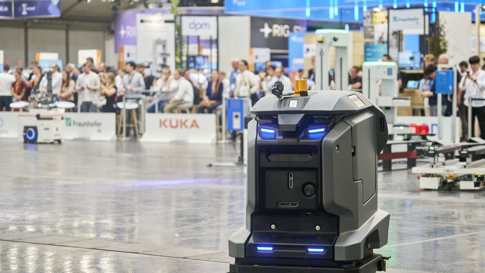 Autonomous mobile robot drives through an exhibition hall, with visitors and exhibitor stands in the background.