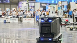Autonomous mobile robot drives through an exhibition hall, with visitors and exhibitor stands in the background.