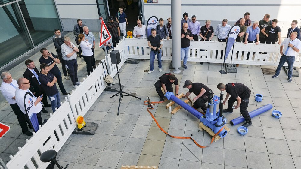 Visitors watch a team of workers assembling a pipe system in front of an exhibition hall, surrounded by safety barriers.