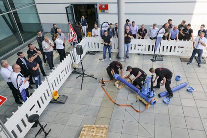 Visitors watch a team of workers assembling a pipe system in front of an exhibition hall, surrounded by safety barriers.