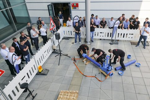 Visitors watch a team of workers assembling a pipe system in front of an exhibition hall, surrounded by safety barriers.