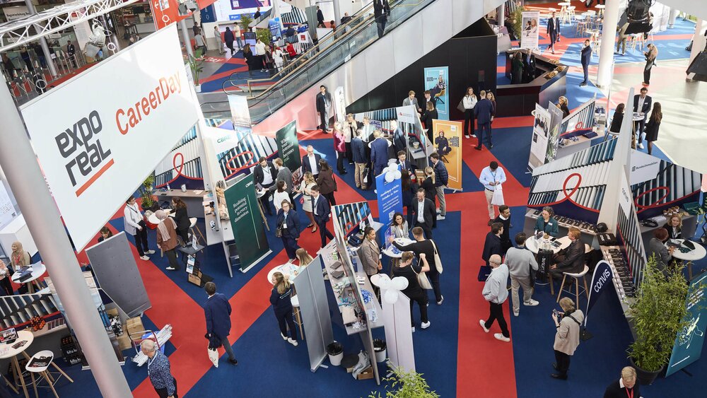Top-down view of the EXPO REAL CareerDay: Many attendees walk between various exhibition booths featuring information displays, counters, and consultation tables. A large banner reading “expo real CareerDay” hangs above the hall. In the background, an escalator leads to another section of the trade fair.