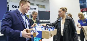 Ein Mann im blauen Anzug hält mehreren Besuchern der BAU einen Karton mit Werbegeschenken entgegen.
