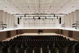 Konzertsaal mit einem Flügel auf der Bühne, leeren schwarzen Stühlen und moderner Beleuchtung an schrägen Wänden und Decke.