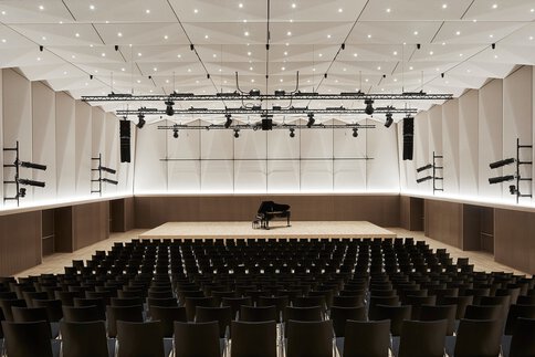 Konzertsaal mit einem Flügel auf der Bühne, leeren schwarzen Stühlen und moderner Beleuchtung an schrägen Wänden und Decke.