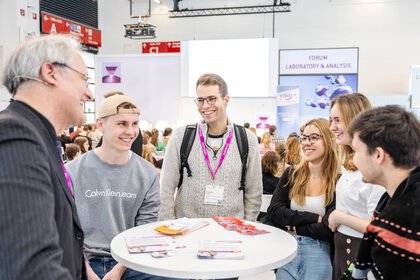 sechs Personen stehen um einen Tisch und führen lachend ein Gespräch