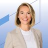 Eine lächelnde Frau mit schulterlangen blonden Haaren und Ohrsteckern trägt eine weiße Bluse und ein beiges Jackett darüber.