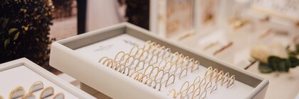 Auf einer Schmucktheke ist eine Ausstellungsschale mit Gold- und Silberringen zu sehen, im Hintergrund sind verschwommene Fotos zum Thema Hochzeit zu erkennen.