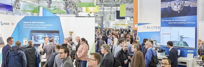 Ein Gang in einer gut besuchten Messehalle der digitalBAU, in dem rechts und links zahlreiche Stände mit Monitoren und bunten Werbebannern stehen.