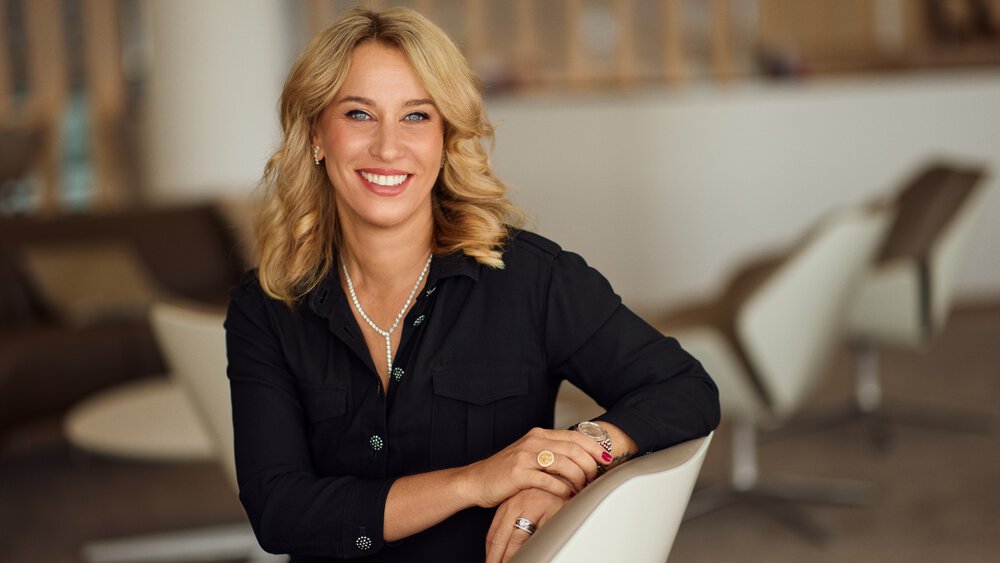 A woman with blonde hair wears various pieces of jewelry and smiles while leaning with one arm on the back of a chair.