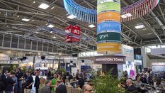 In a well-attended exhibition hall at transport logistic, there is a circular snack bar with a colorful pillar with logos and the inscription 