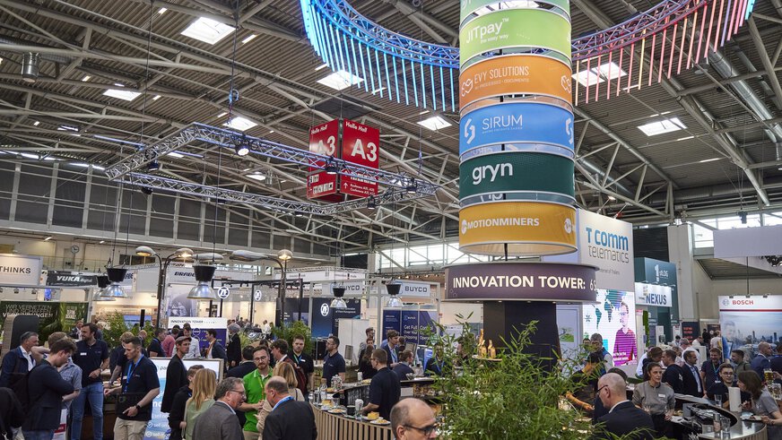 In a well-attended exhibition hall at transport logistic, there is a circular snack bar with a colorful pillar with logos and the inscription 