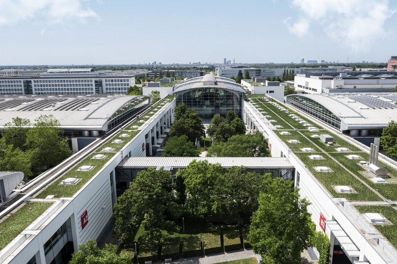 A view from above of the partially greened hall roofs and courtyards on the Messe München site. The city’s skyline can be seen in the background.