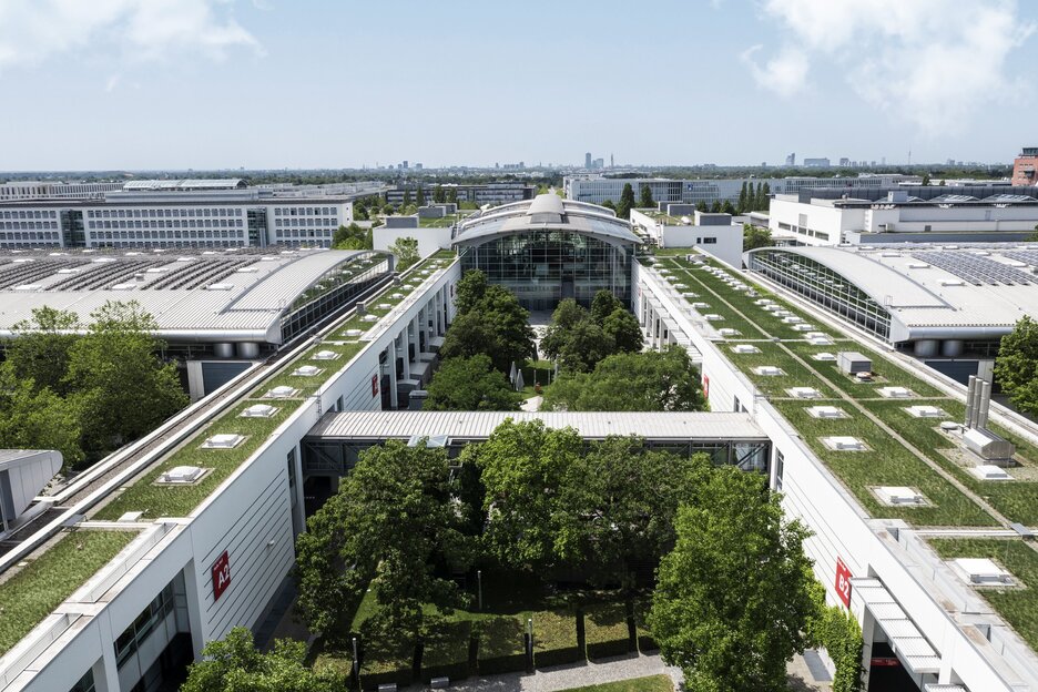 A view from above of the partially greened hall roofs and courtyards on the Messe München site. The city’s skyline can be seen in the background.