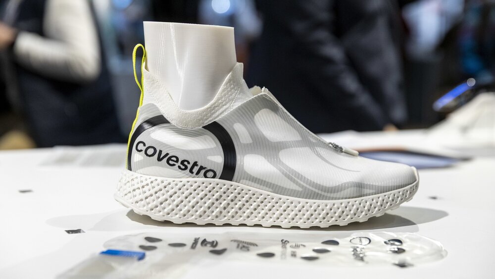 Nahaufnahme eines weißen und grauen Sportschuhs mit der Aufschrift „covestro“ mit einem Puppenfuß darin auf einer Präsentationsfläche der LOPEC.