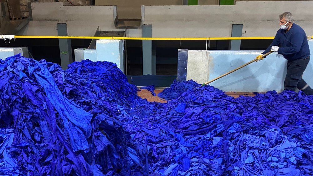 Große Mengen knallblauer Textilreste liegen in einer Produktionshalle, während eine Person mit einem langen Werkzeug das Material bewegt.