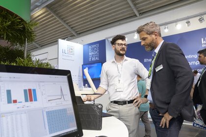 Zwei Businessmänner unterhalten sich an einem Messestand über ein Exponat bestehend aus einem Bildschirm mit verschiedenen Diagrammen darauf