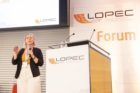 Eine Frau mit einem Mikrofon in der Hand steht neben einem Podium im LOPEC Forum und spricht zu einem Publikum außerhalb des Bildes.