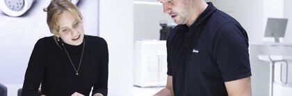 Two people at an exhibition stand bend over a light table on which various technical components are lying.